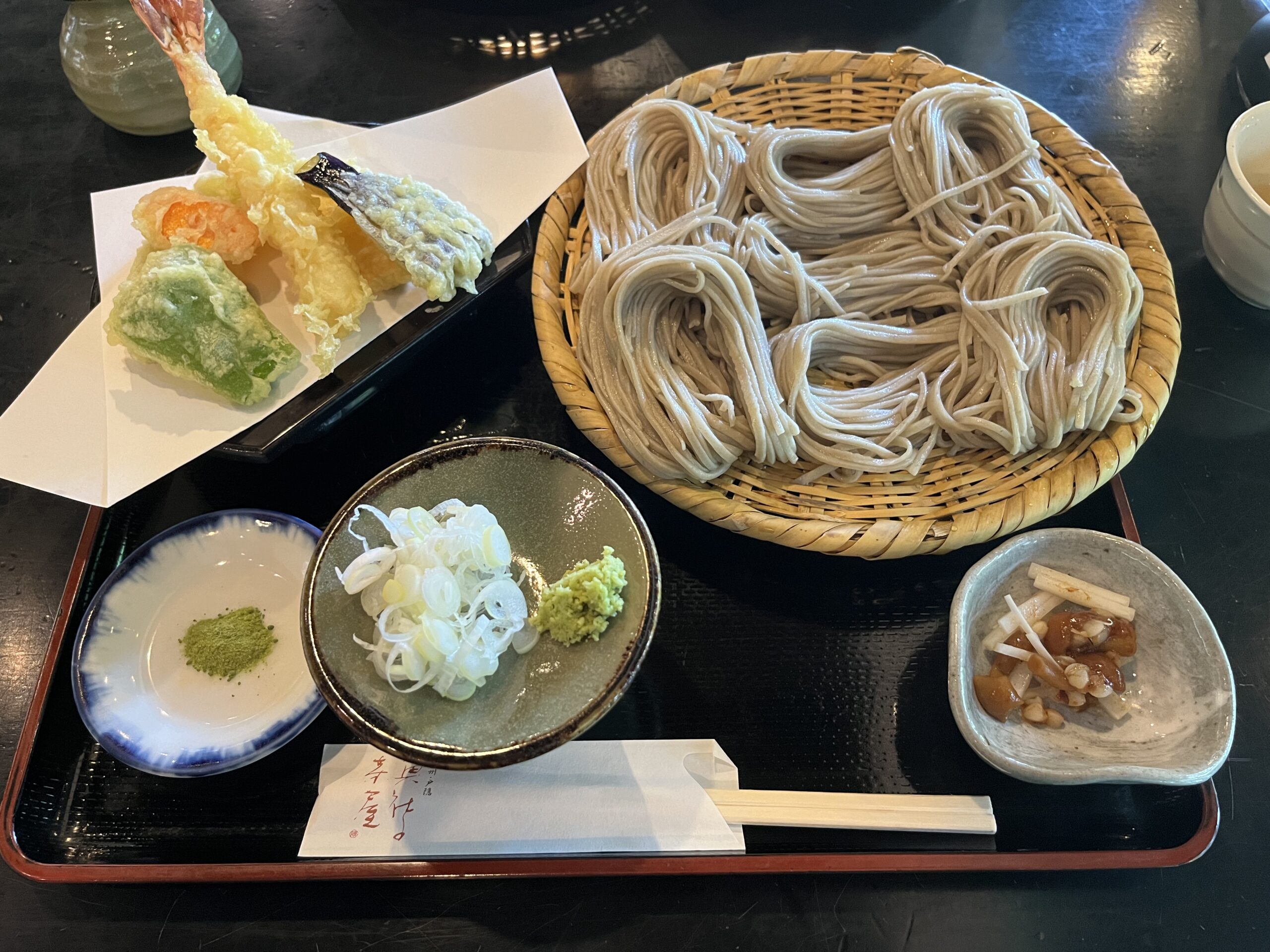 Zaru soba noodles in a bamboo basket on a black tray with tempura, grated wasabi, chopped scallions and a small side dish