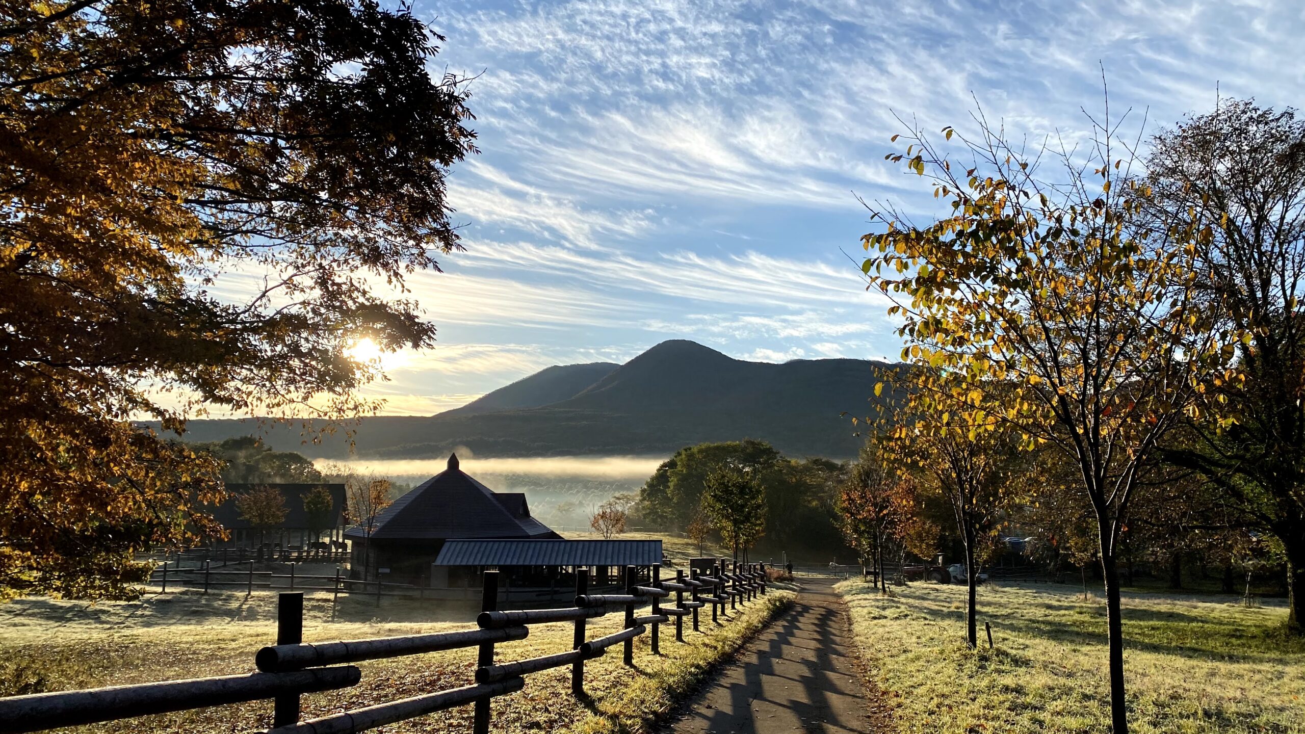 Frosty meadow path and wooden fence in autumn morning light with a small building and a mountain in the background